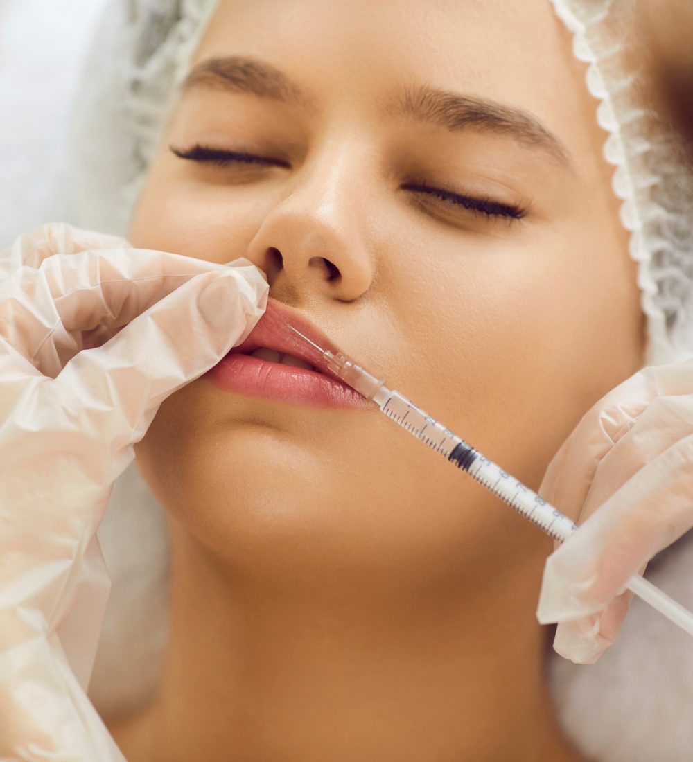 Beautiful woman getting professional treatment at clinic of aesthetic medicine. Young lady receiving botox injection for bigger, plumper lips. Doctor's hands holding syringe above face in close up